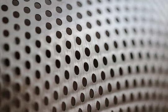 Close-up on a metallic perforated surface or metal grate with uniformly sized black circular holes, creating a pattern and an industrial texture
