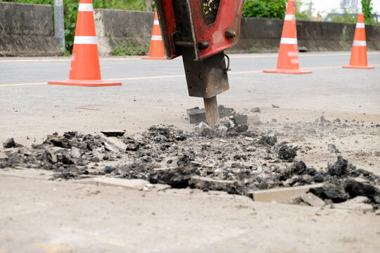 Construction workers are repairing a road by excavating and drilling the pavement to fill underground voids with specially mixed concrete. Heavy machinery and roadwork safety cones are visible.