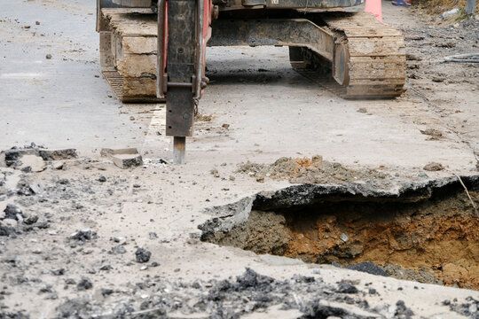 Construction workers are repairing a road by excavating and drilling the pavement to fill underground voids with specially mixed concrete. Heavy machinery and roadwork safety cones are visible.