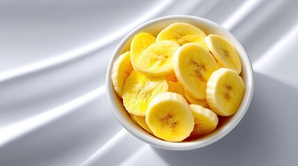 Top Down View of a White Bowl Filled with Sliced Yellow Bananas on a Clean White Surface with Dramatic Lighting Creating Shadows