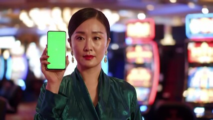 Elegant Asian Woman Holding a Green Screen Smartphone in a Casino.