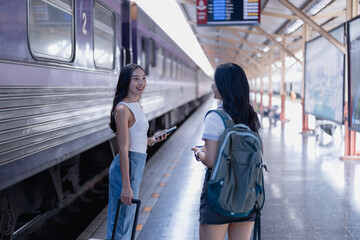 Obraz premium Two young adult women friends standing on a train station platform, talking and smiling while preparing to travel by train, with luggage and a backpack