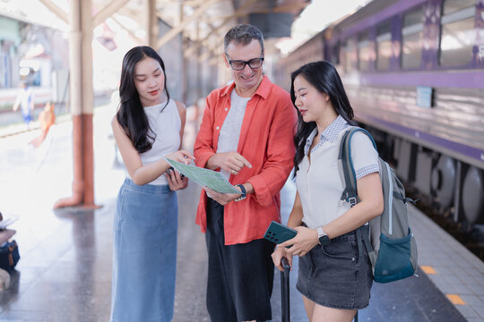 Three diverse friends, including two women and one man, are standing on a train station platform, actively discussing their travel plans and pointing at a physical map, preparing for their journey