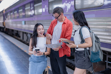 Diverse group of three friends, two young women and a senior man, standing on a train station platform, smiling and looking at a map while planning their travel adventure together