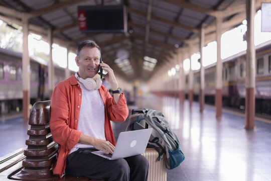 Traveler man sitting on a bench at a railway platform, talking on his smartphone and using a laptop while waiting for transport, embodying remote work and travel lifestyle