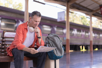 Mature man sits on a train platform bench studying a paper map, backpack and luggage nearby as he...