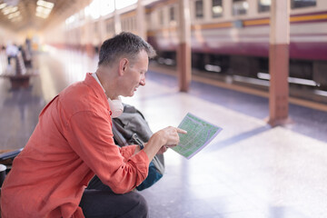 Mature man sitting on a train platform, holding a physical map and pointing at a location, planning...