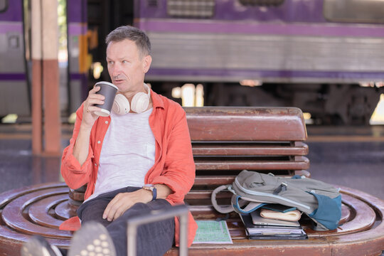 Mature man sits on a wooden bench at a train platform, sipping takeaway coffee with headphones around his neck and a backpack by his side, waiting thoughtfully for his train