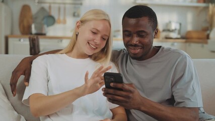 Carefree lovers watching funny content in social media on smartphone display. Medium portrait of happy interracial spouses in apartment, internet addiction and mobile gadget overuse of modern people