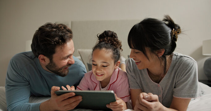 Parents, girl and tablet in bed, smile and bonding with online games, scroll or problem solving in home. People, mother and daughter with dad, tech and relax with app for puzzle at family house - Powered by Adobe