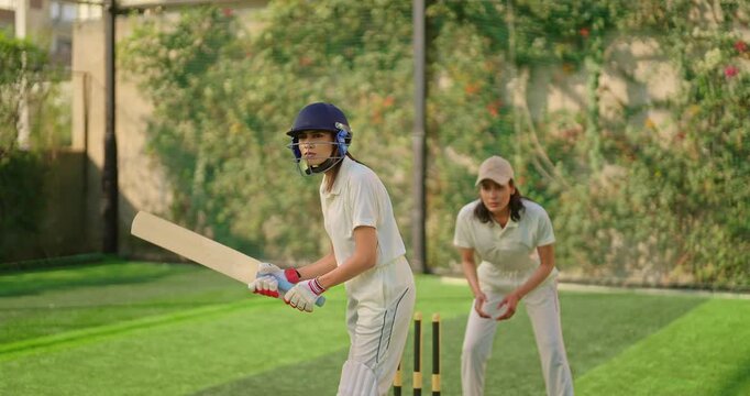 Indian gen z young adult woman lady wear hand gloves white dress leg guard helmet day time cricket player sports teen female girl hold bat play casual match game hit ball do batting at ground place	
