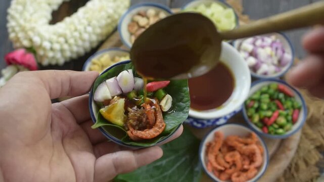 This video shows a person pouring sweet sauce onto a traditional Thai snack called Miang Kham, which consists of betel leaves filled with roasted coconut, dried shrimp, lime, shallot, chili.
