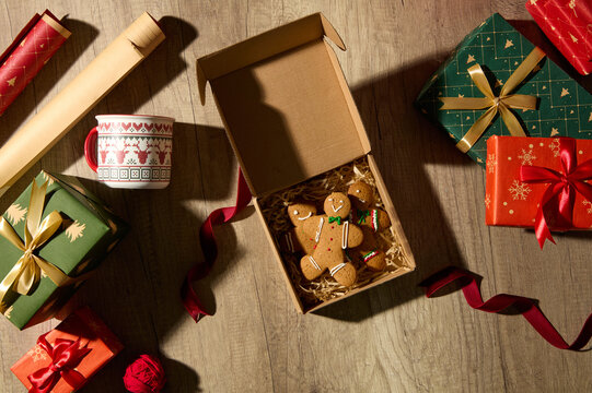 Christmas flat lay with cookie gift box, green and red wrapping paper, ribbons, and golden decorations on wooden surface. Warm handmade festive concept.
