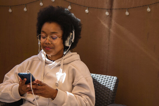 African American woman relaxing on the terrace of her house listening to music with headphones - Powered by Adobe