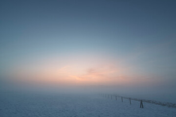 Peaceful winter sunrise over snow-covered fields with soft fog and pastel sky