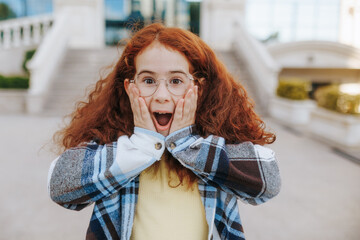 Surprised redhead teenage girl with glasses showing shocked OMG expression outdoors