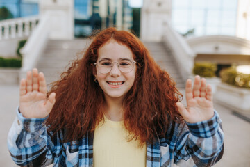 Smiling redhead teenage girl with glasses waving hello outdoors in casual outfit