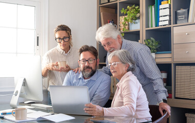 Group of mature and senior professionals working together in a modern office. Men and women collaborate around a laptop discussing ideas and reviewing information. Teamwork, mentoring, cooperation