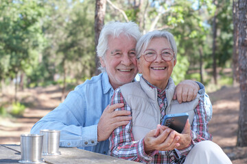 Happy senior couple enjoying a picnic outdoors in a sunny forest using smartphone. Elderly man and woman sitting at a wooden table smiling sharing social media on phone. Positive lifestyle in nature