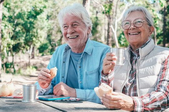 Happy senior couple enjoying a picnic outdoors in a sunny forest. The elderly man and woman are sitting at a wooden table, smiling sharing food and drinks. Positive retired lifestyle in nature