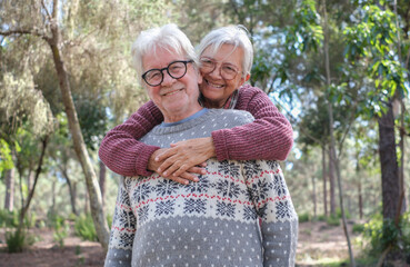 Happy senior couple enjoying a moment of affection and emotion outdoors in a sunny forest. The elderly woman lovingly hugs her partner from behind while both smile warmly at the camera