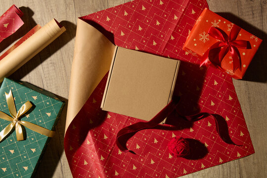 Christmas packaging mockup with kraft box, red wrapping paper, and ribbons on wooden surface. Warm handmade mood perfect for holiday branding or product scene.