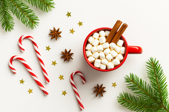 Flat lay of red mug with hot chocolate and marshmallows, candy canes, cinnamon sticks, and fir branches on white background. Festive Christmas composition with holiday mood and copy space.