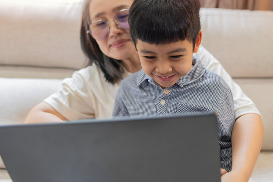 Cheerful Asian mother and cute son laughing while sitting on sofa with laptop at home.
