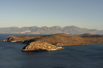 View of the historic Spinalonga Island fortress surrounded by the turquoise waters of the Aegean Sea near Elounda, Crete, Greece. The image captures the dry rocky landscape, ancient stone walls