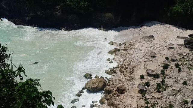 Waves crash softly against the rocky shore of Sawangan Beach, Kebumen. The coarse sandy beach, mixed with pebbles and coral, blends beautifully with large limestone rocks and gentle sea foam.