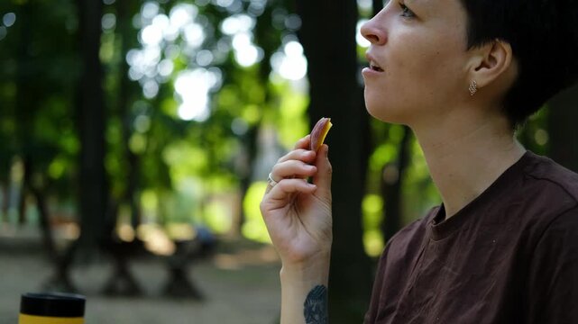 Woman with short hair enjoys eating a slice of ripe peach while sitting in green park on a sunny summer day, relaxed and thoughtful. 4k horizontal footage