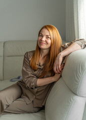 Obraz premium Cute young woman sitting on sofa at home, smiling. Relaxed lifestyle, cozy indoor moment, happy and cheerful, casual home comfort