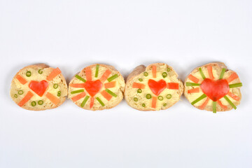 Four baguette slices with butter are decorated with tomato hearts, tomato slices, and sliced scallions, arranged in a horizontal line on a white surface