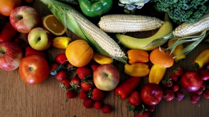 A top down view slowly pans over a colorful assortment of fresh fruits and vegetables, including apples, corn, peppers, and bananas, arranged on a wooden table. - Powered by Adobe