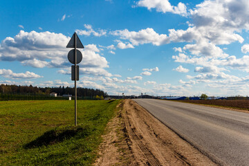 Back of a road sign on a country roadside, flanked by a lush green field and an open asphalt road under a bright blue sky with fluffy cumulus clouds
