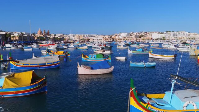 Aerial view of Marsaxlokk harbour in Malta with luzzu boats and skiffs on deep blue water. The parish church with twin bell towers rises behind in crisp daylight.