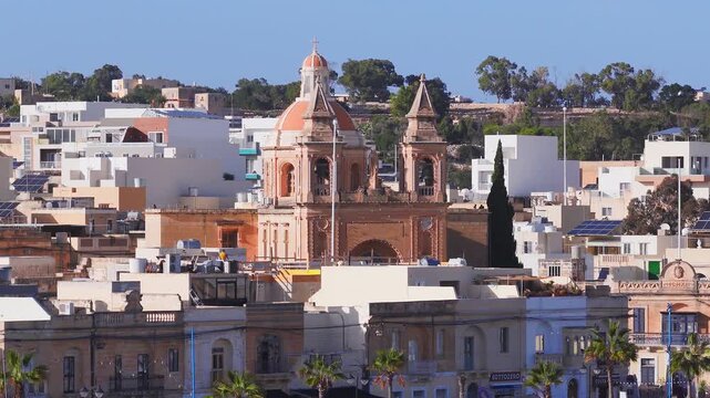 Aerial pan across Marsaxlokk, Malta, shows a sandstone parish church, orange dome, twin towers, low flat roofs with solar panels, palm trees, workers, and clear daytime light.