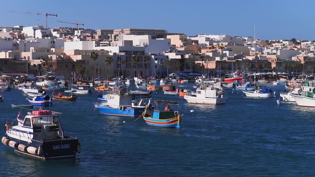 Aerial view shows Marsaxlokk harbor in Malta, luzzu and workboats bob on blue water, sunlit limestone quay with palms and awnings, cranes and new blocks behind.