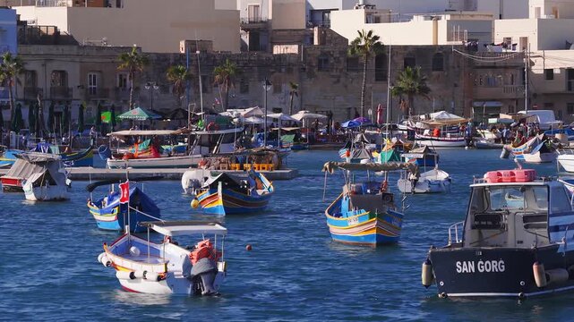 Aerial view of Marsaxlokk harbor in Malta shows luzzus, the SAN GORG boat, palm lined quay, visitors, fishermen, market stalls, and a boat trips sign at midday.