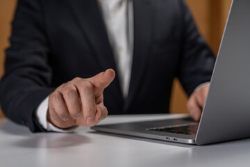 Businessman in suit touching invisible screen or interface while working on laptop at desk in modern office environment, showing technology interaction concept.