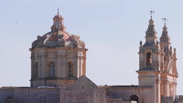 Aerial pan across Mdina Cathedral in Malta shows dome and bell towers, pink honey limestone, cartouches, volutes, weathervanes, clear daylight, tight framing.