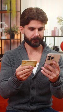 Indian man at home holds smartphone and bank card enters cvv completes payment gets confirmation. Arabian guy on sofa shows approval hand sign nods yes smiles after online purchase verified order paid