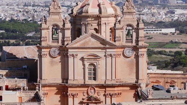 A slow aerial pan centers St Pauls Cathedral in Mdina, Malta, with twin bell towers, clock faces, and pink dome, revealing fields and a modern urban belt across the valley.