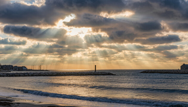 Lighthouse on breakwater with sunbeams through clouds over ocean.