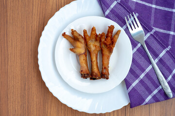 Description: A high-angle, top-down shot showing three deep-fried chicken feet, a food rich in collagen, served on a white plate over a purple napkin and wooden table.