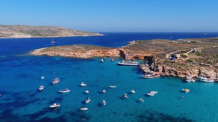 Aerial panorama shows Blue Lagoon on Comino Island, Malta. Boats and a tour vessel rest in turquoise water as swimmers and sunbathers gather under midday light.