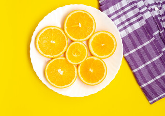 A top-down view shows several round slices of orange fruit (Citrus × sinensis) on a white plate. The plate is on a bright yellow surface next to a purple checkered cloth.