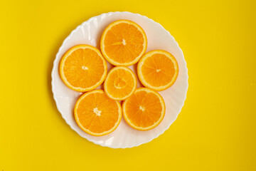 A top-down view shows several round slices of orange fruit (Citrus × sinensis) arranged on a white plate, which is set on a solid, bright yellow background with copy space.