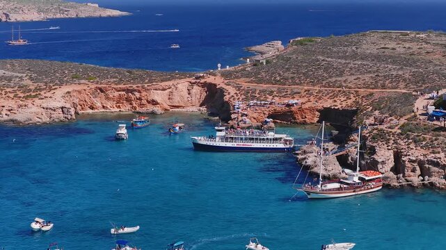 Aerial view of Comino Blue Lagoon, Malta. Hornblower, yachts, and dinghies cruise and moor. Swimmers, umbrellas, red cliffs, and turquoise water define summer scene.