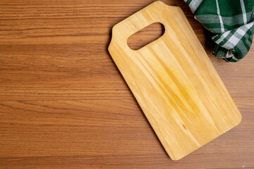 light wooden cutting board with a handle cutout rests on a dark wooden table, viewed from above. A green and white checkered cloth is in the upper right corner.